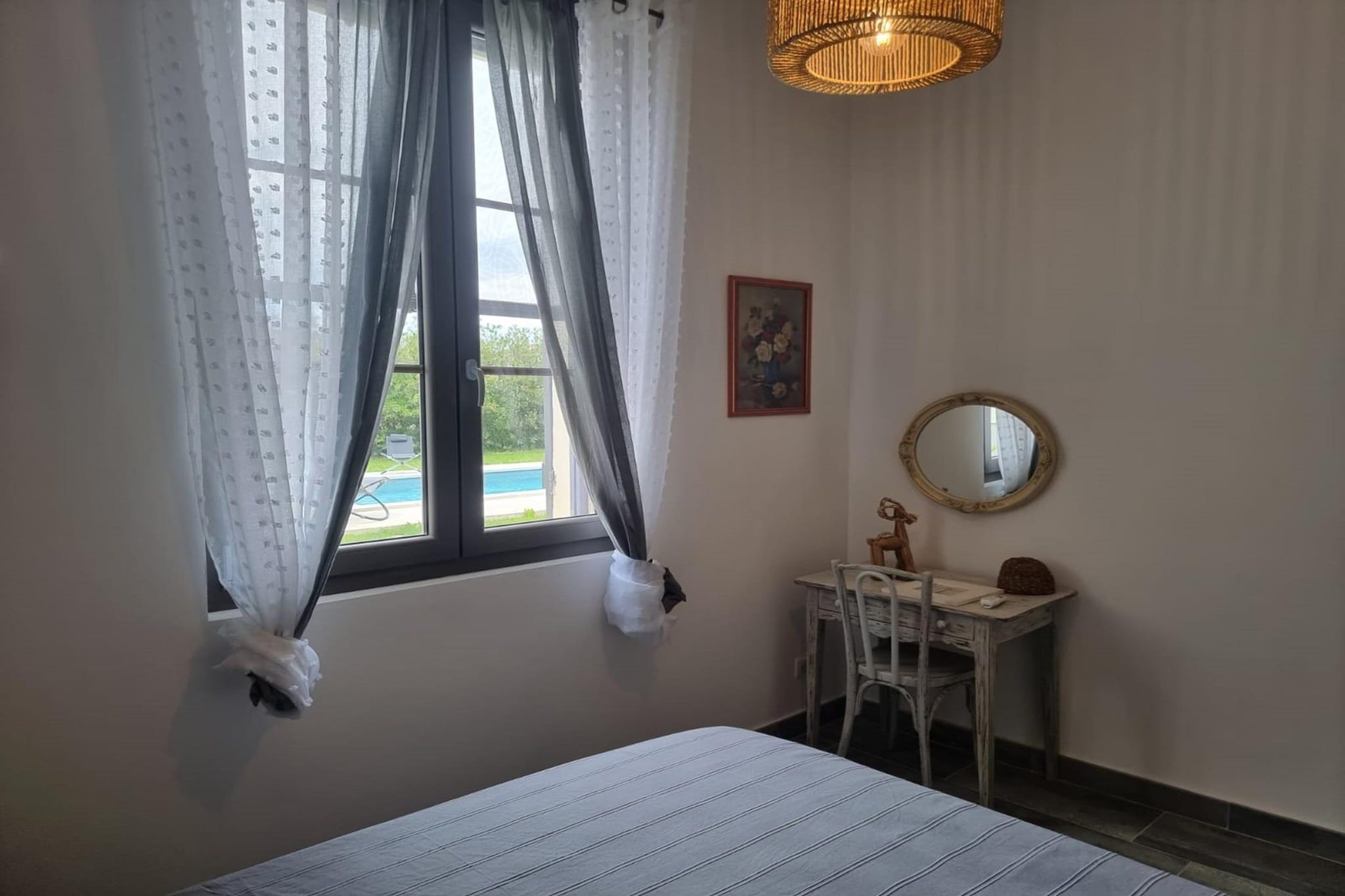 A bedroom with a bed, a desk, a chair, a window with curtains, a hanging light fixture, and a wall-mounted mirror.