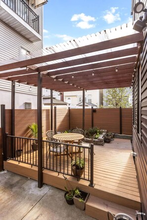 Une terrasse couverte avec une table et des chaises en bois, entourée de plantes en pot et d’une rambarde en métal.