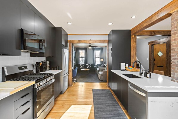A modern kitchen with stainless steel appliances, a wooden ceiling, and a view into a living area.