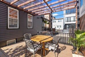 Une terrasse couverte avec une table ronde et des chaises, un petit jardin avec des plantes et une vue sur les bâtiments voisins.
