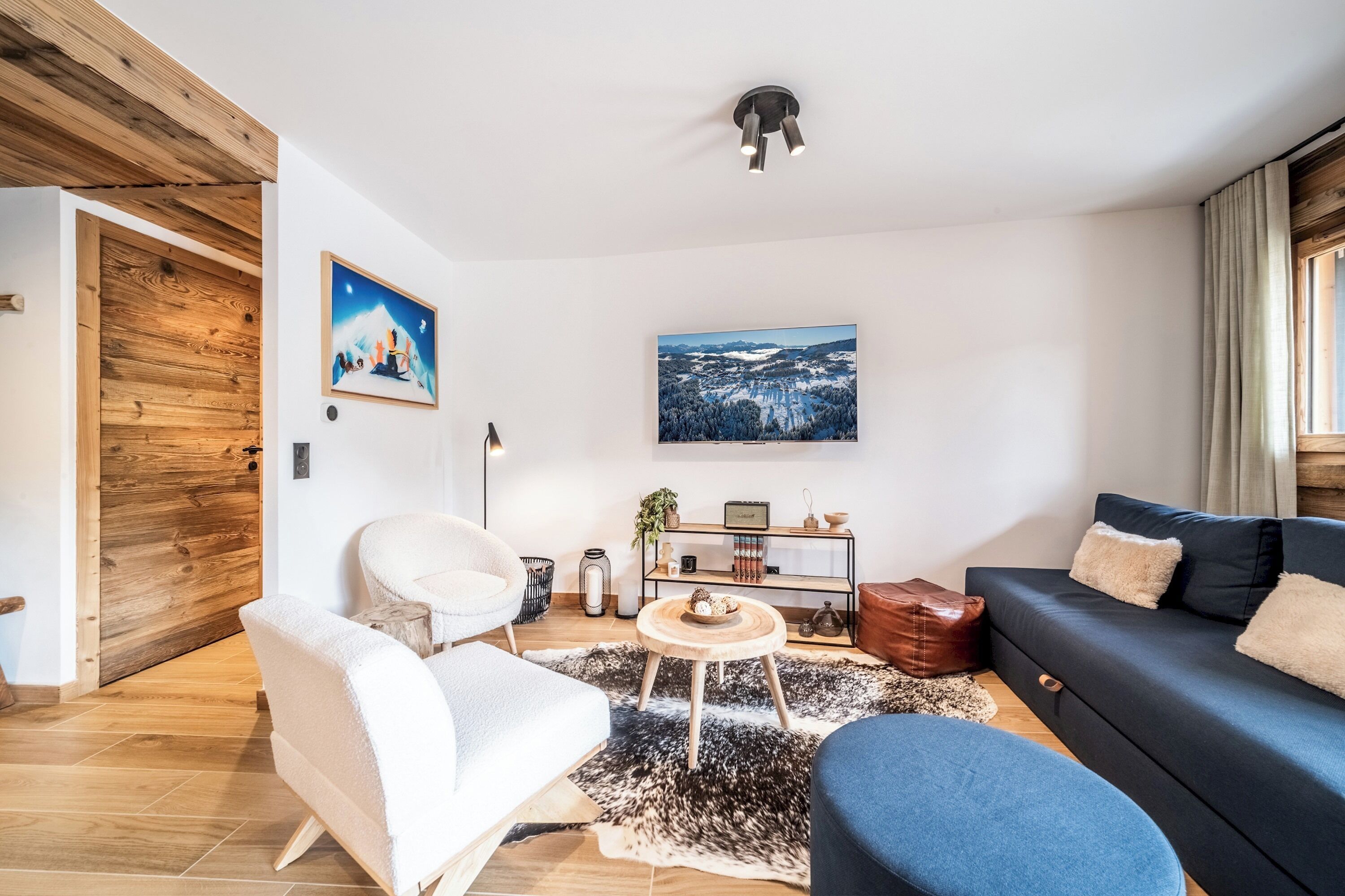 Ein modernes Wohnzimmer mit Holzboden, einem schwarzen Sofa, einem weißen Sessel, einem blauen Hocker, einem runden Couchtisch und einem schwarz-weißen Teppich.