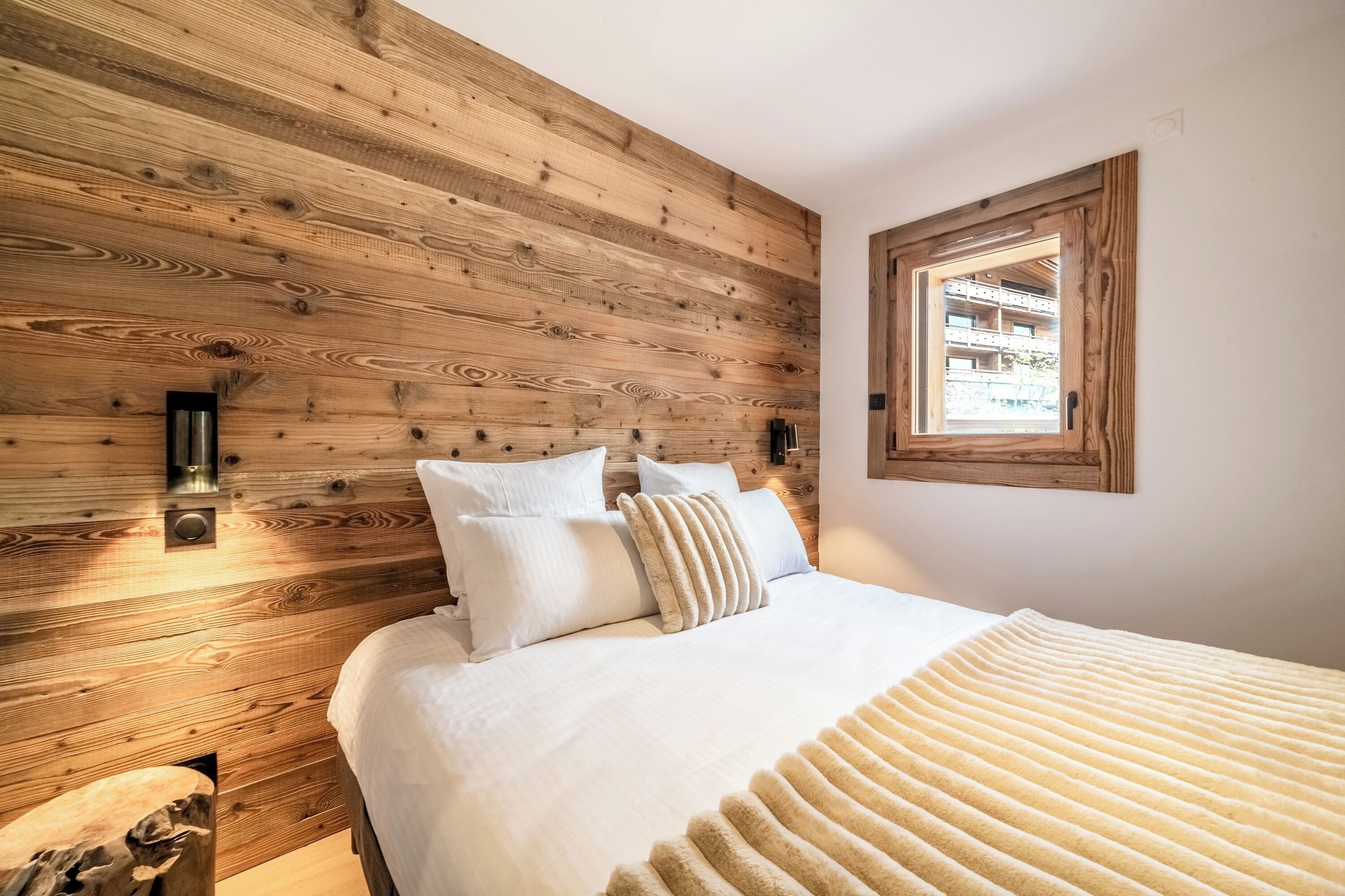 A bedroom with a wooden wall, a bed with white bedding, and a window with a view of buildings.