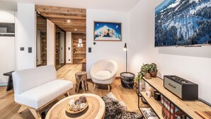 Ein modernes Wohnzimmer mit einem weißen Sofa, einem Holztisch, einem weißen Sessel und einem Bücherregal mit Büchern und einem Lautsprecher.