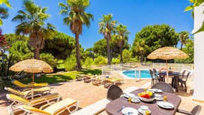 A poolside area with lounge chairs, a dining table set for two, and a pool surrounded by palm trees and greenery. - Villa Mexico - Four Bedroom Villa, Sleeps 8 (Quarteira)