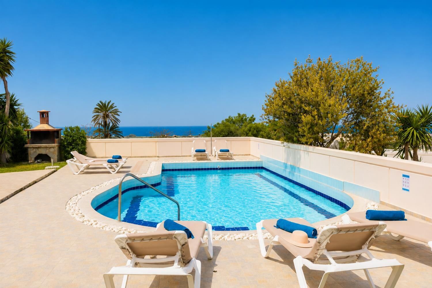 Una piscina rectangular con borde azul, rodeada de sillas de descanso con cojines azules, una chimenea de piedra y vistas al mar.