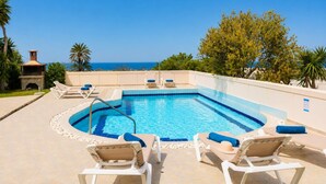 A rectangular swimming pool with a blue border, surrounded by lounge chairs with blue cushions, a stone fireplace, and a view of the sea. - Villa Maia - Three Bedroom Villa, Sleeps 6 (Rhodes)
