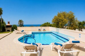 Una piscina rectangular con borde azul, rodeada de sillas de descanso con cojines azules, una chimenea de piedra y vistas al mar.