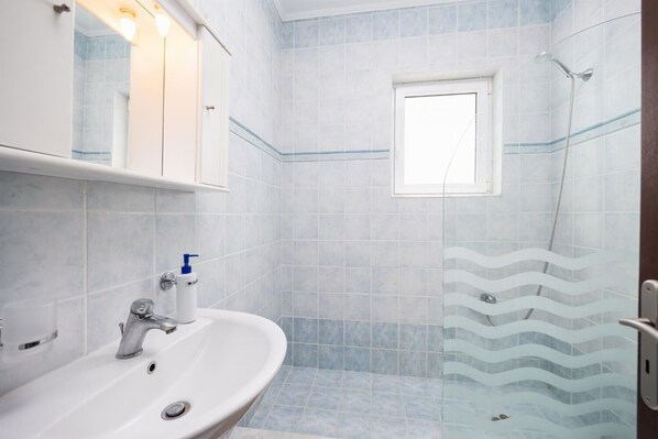 Une salle de bain avec un lavabo blanc, un miroir, une douche avec une porte en verre et une petite fenêtre.