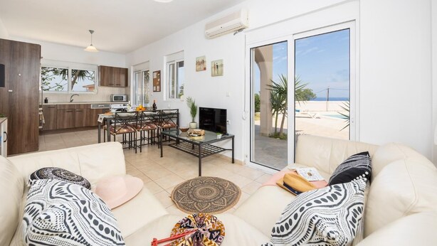 A spacious living room with a sofa, coffee table, and a TV. There is a kitchen area with wooden cabinets and a dining table. The room has large sliding glass doors leading to a balcony with a view of the beach. - Villa Maia - Three Bedroom Villa, Sleeps 6 (Rhodes)