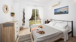 A bedroom with a bed, a dresser, a window with curtains, a wall-mounted air conditioner, and a painting. - Villa Maia - Three Bedroom Villa, Sleeps 6 (Rhodes)