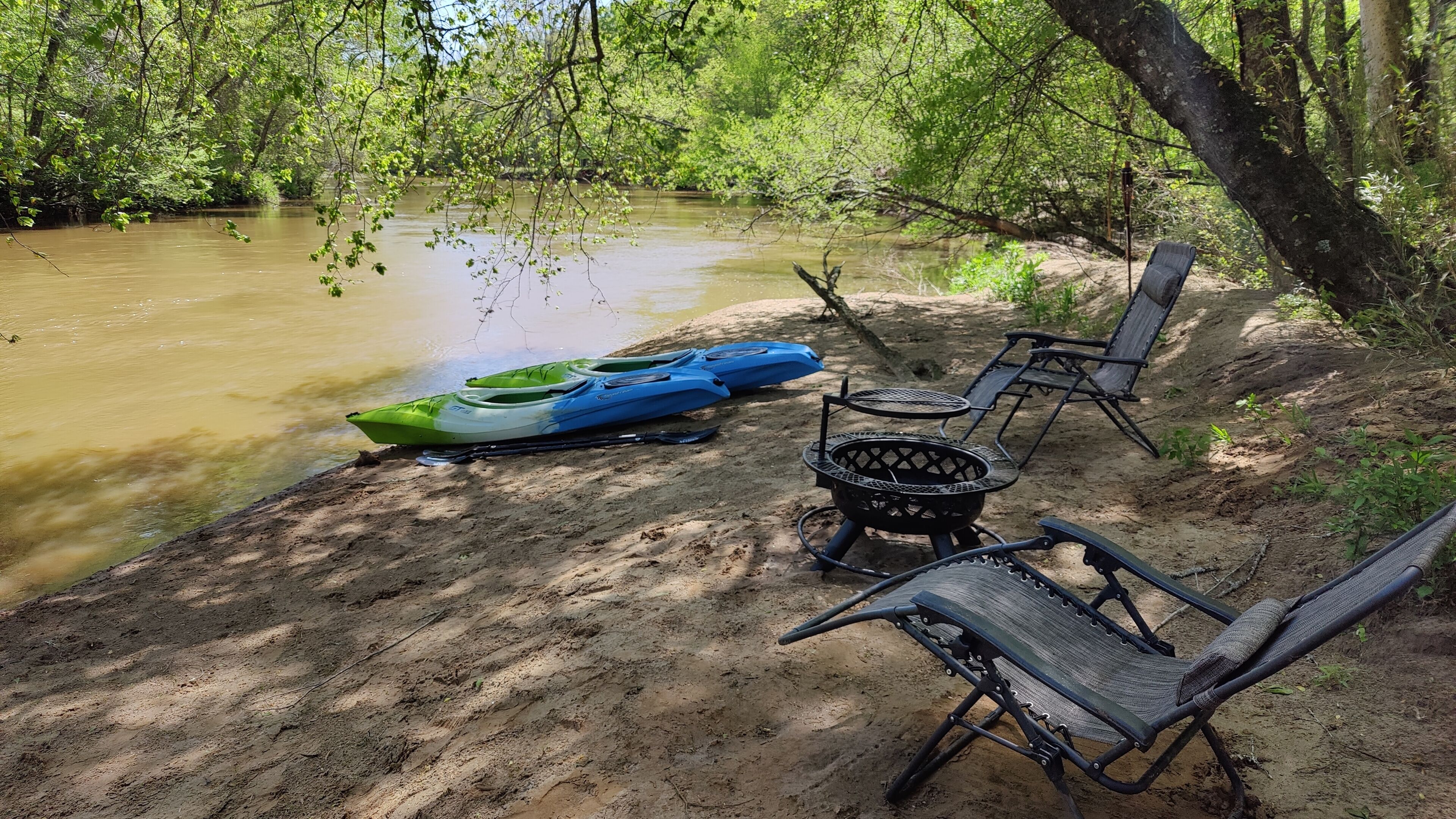 Un terrain de camping au bord de la rivière, équipé de kayaks, d’une table et de chaises.