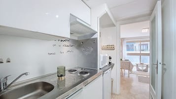 Eine kompakte Küche mit Spüle aus Edelstahl, einer Mikrowelle und einem Herd. An der Wand hängt ein handgeschriebener Zettel und über der Spüle befindet sich ein kleines Fenster.