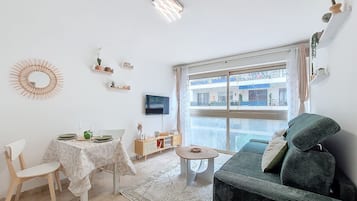 Ein Wohnzimmer mit einer Couch, einem Esstisch mit Stühlen, einem an der Wand befestigten Fernseher und einem großen Fenster mit Blick auf Gebäude.