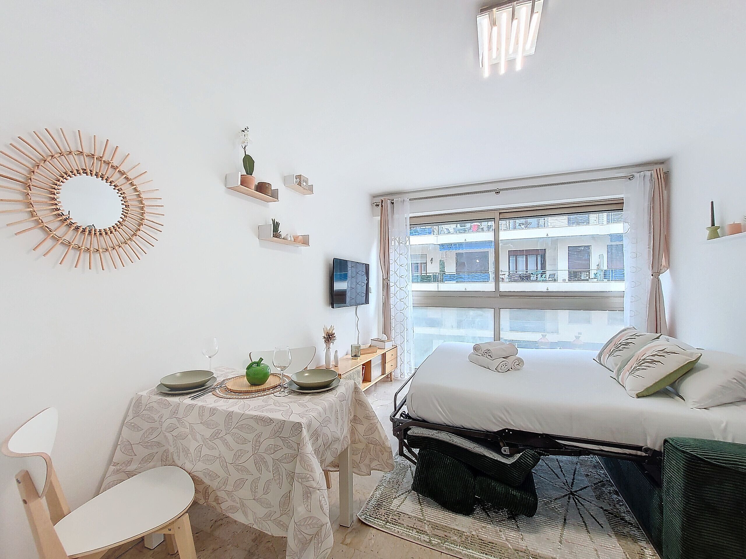 Une chambre à coucher avec un lit, un bureau avec une table, une chaise, une télévision et une fenêtre donnant sur des immeubles.