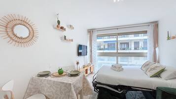 Une chambre à coucher avec un lit, un bureau avec une table, une chaise, une télévision et une fenêtre donnant sur des immeubles.