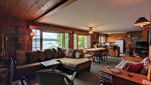 A cozy cabin interior with a sofa, dining table, kitchen area, and large windows overlooking a lake. - Winnipesaukee Heirloom Cottage (Meredith)