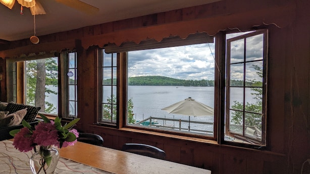 A wooden cabin with large windows overlooking a lake, a table with a vase of flowers, and a chair. - Winnipesaukee Heirloom Cottage (Meredith)