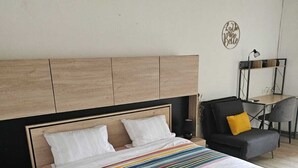 A bedroom with a wooden headboard, a colorful striped bedspread, a black chair with a yellow pillow, and a desk with a lamp.