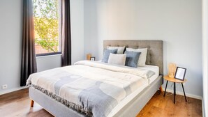 A modern bedroom with a large bed, a window with curtains, a bedside table with a lamp, and a view of greenery outside. - Charming Ostend Home: Near Beach & City Center (Oostende)