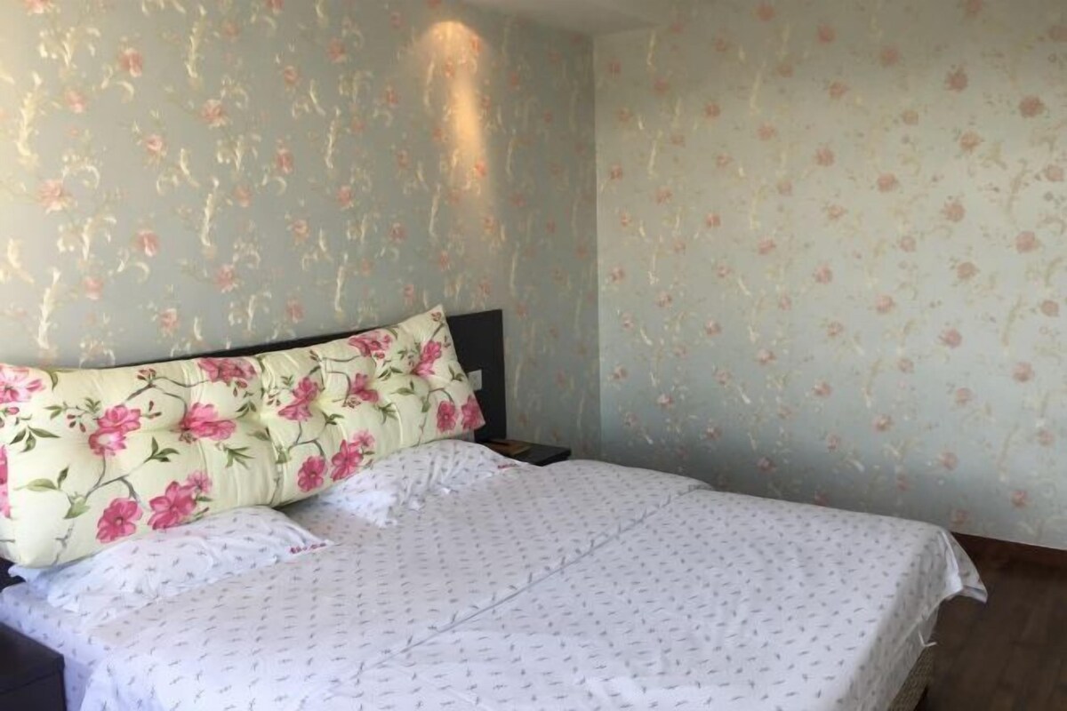 Une chambre à coucher avec un papier peint à motifs floraux, un lit recouvert d’une courtepointe blanche et une table de chevet avec une lampe.