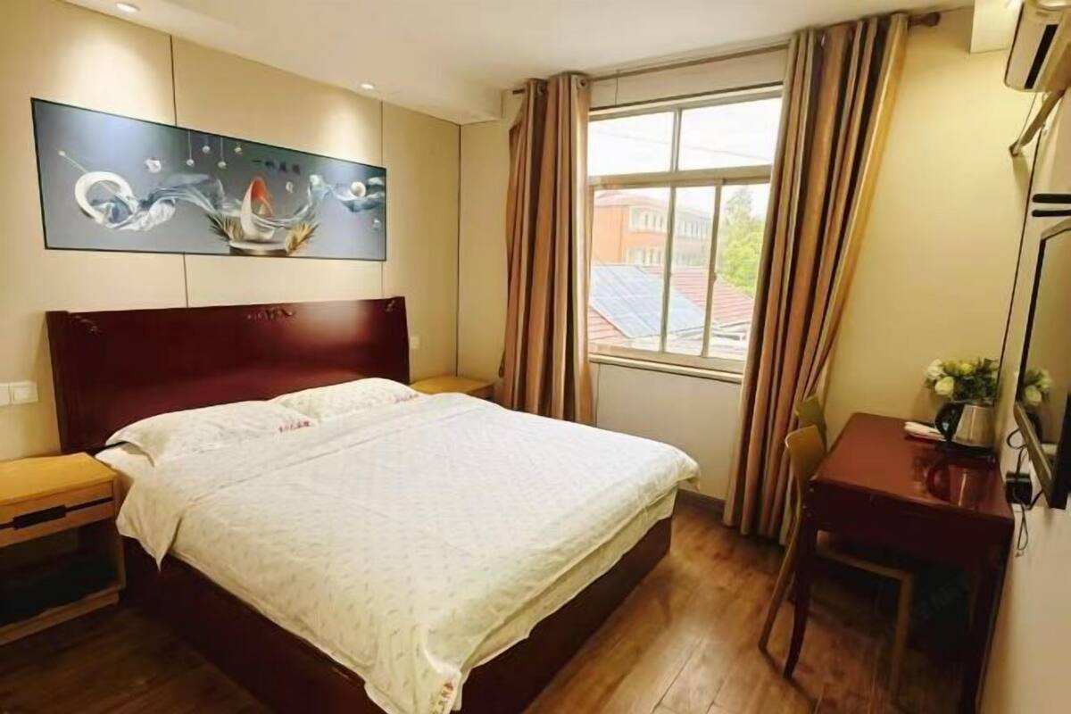 A hotel room with a large bed, a desk, a chair, a window with curtains, and a wall-mounted artwork.