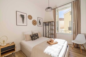 Ein Schlafzimmer mit Bett, Nachttisch mit Lampe, Stuhl, Fenster mit Vorhängen und Wandbild.
