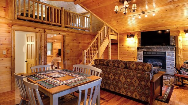 A wooden cabin interior with a dining area, sofa, fireplace, and a staircase leading to a loft. - Cozy Cabin with second floor loft, outdoor BBQs, campfire pits, and more! (Lake Placid)