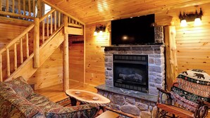 A log cabin interior with a stone fireplace, wooden staircase, and cozy seating area. - Cozy Cabin with second floor loft, outdoor BBQs, campfire pits, and more! (Lake Placid)