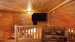 A wooden cabin interior with a television mounted on the ceiling, a wooden staircase, and a log bench. - Cozy Cabin with second floor loft, outdoor BBQs, campfire pits, and more! (Lake Placid)