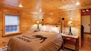 A cozy log cabin bedroom with a bed, two bedside tables with lamps, a window, and a door. - Cozy Cabin with second floor loft, outdoor BBQs, campfire pits, and more! (Lake Placid)