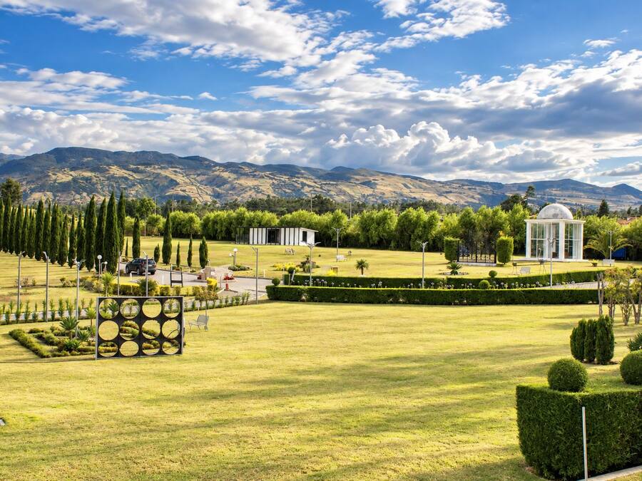 Um jardim bem cuidado com gramados aparados, um gazebo e uma fileira de ciprestes.