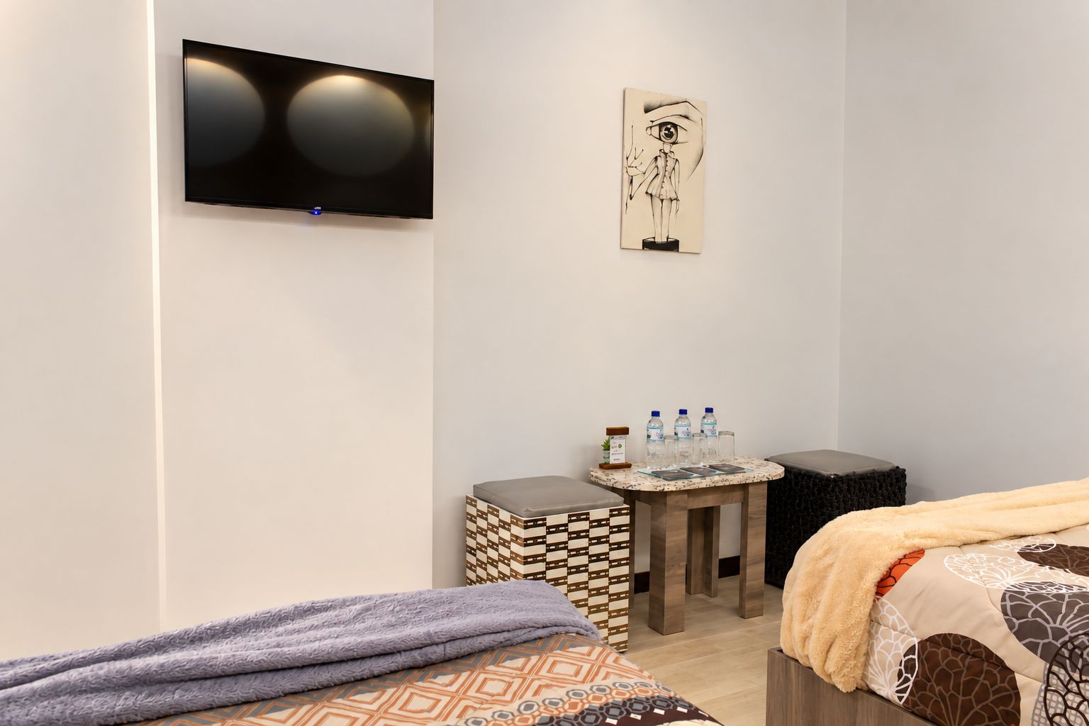 A hotel room with a bed, a television mounted on the wall, a small table with water bottles, and a decorative piece above the table.