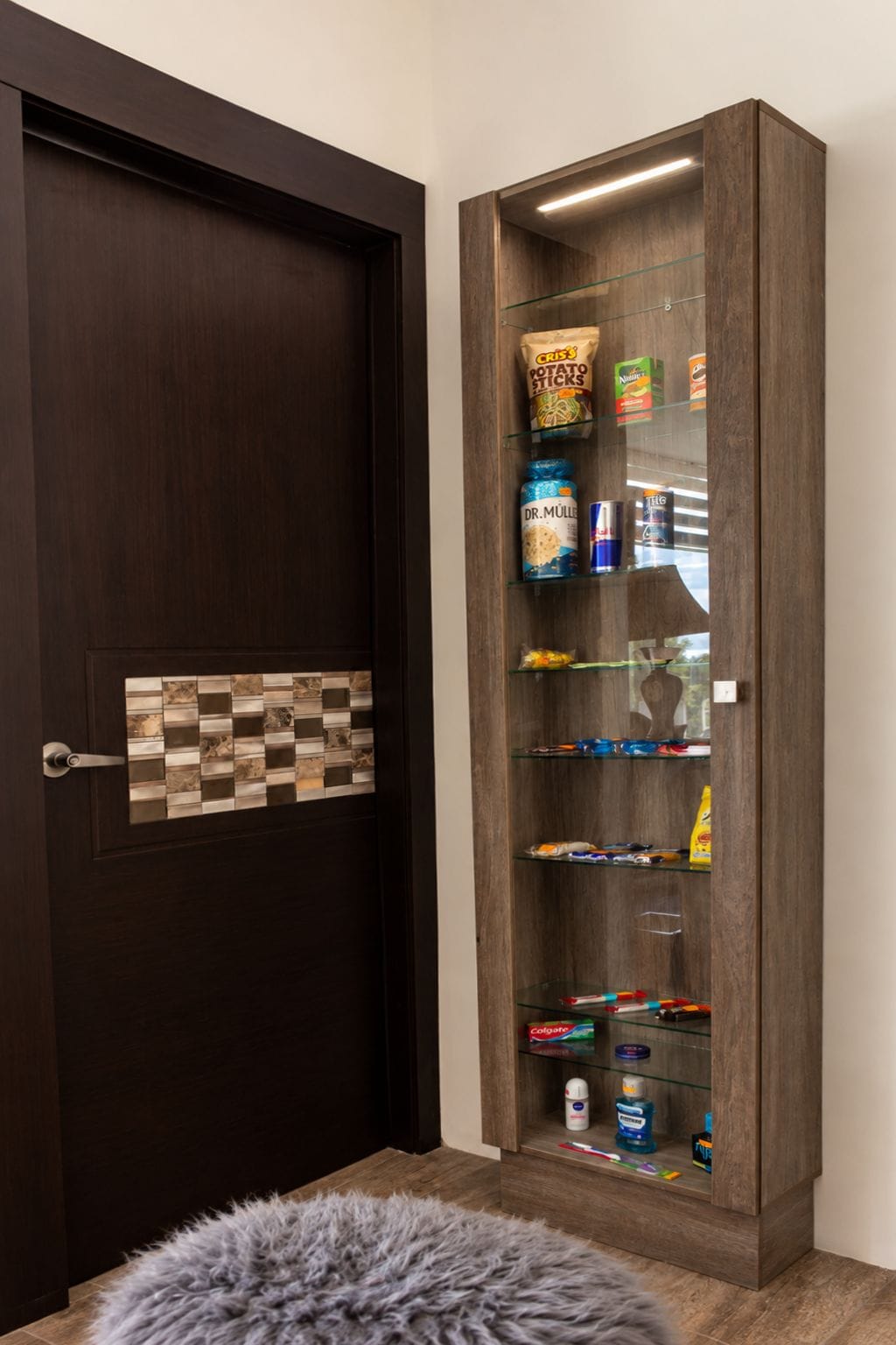 Una puerta de madera oscura con un diseño de baldosas en tablero de ajedrez. A la derecha, un mueble con puertas de cristal y estantes que contienen diversos artículos como aperitivos, bebidas y productos de higiene personal.