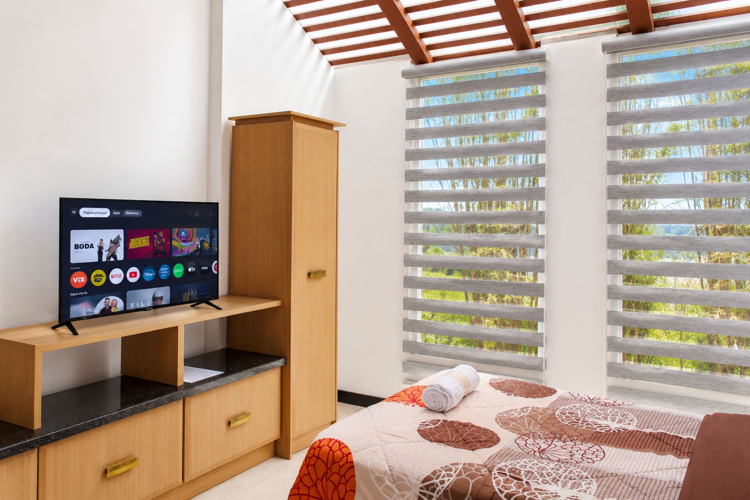 Un dormitorio con cama, televisor sobre un mueble de madera, ventana con persianas y vista a zonas verdes.