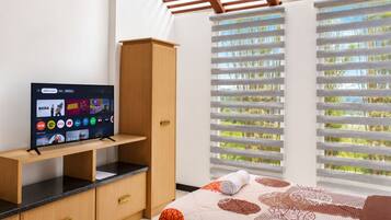 Un dormitorio con cama, televisor sobre un mueble de madera, ventana con persianas y vista a zonas verdes.