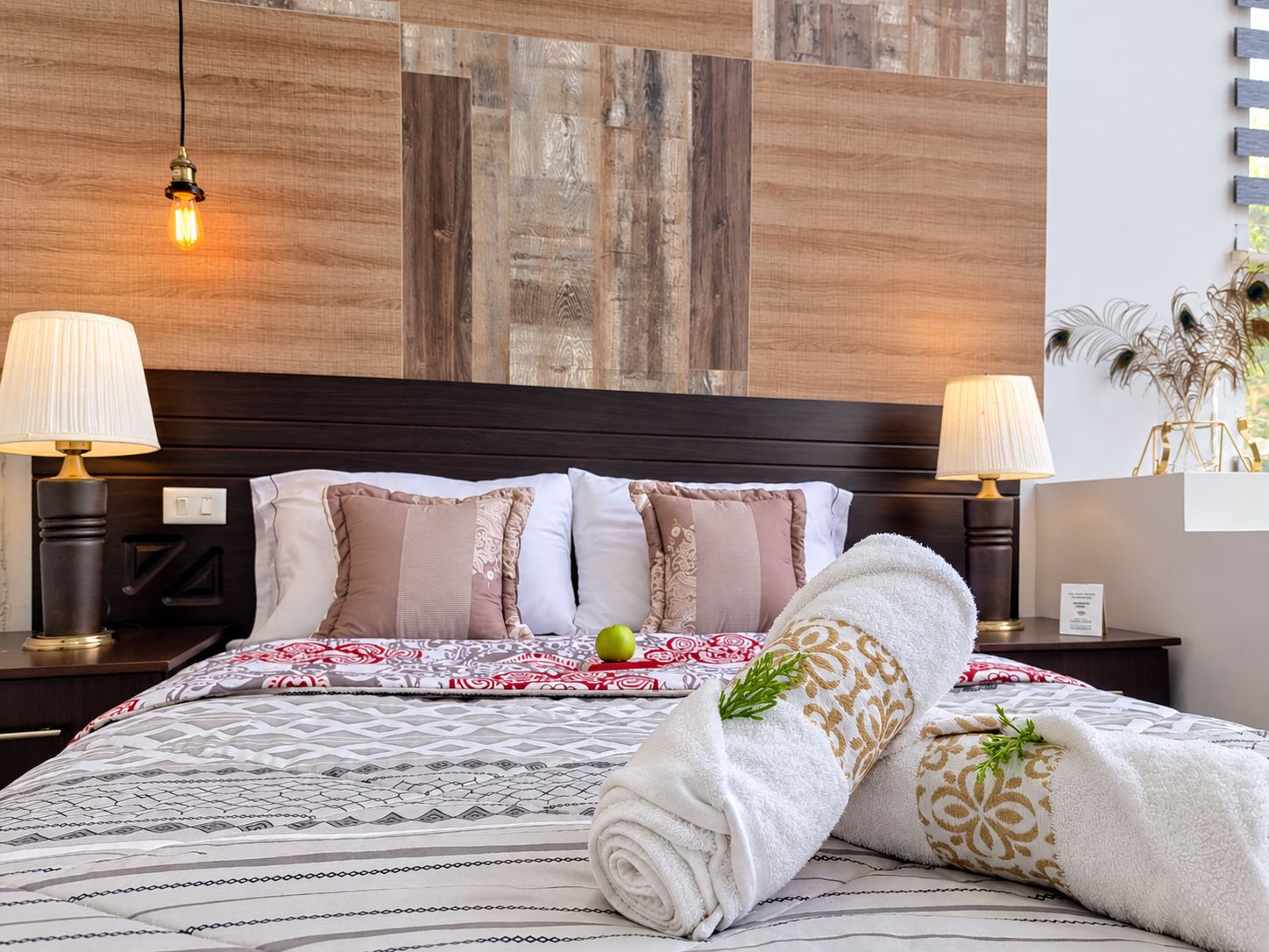 Una habitación de hotel con cabecera de madera, dos lámparas de noche, una colcha estampada y una toalla enrollada sobre la cama.