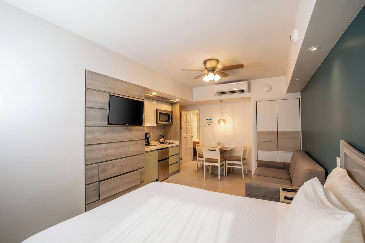 A modern hotel room with a large bed, a dining area, a kitchenette, and a ceiling fan.