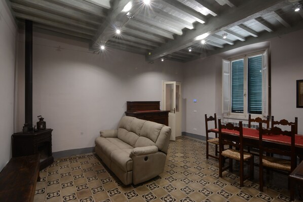 A room with a sofa, dining table, and chairs, wooden floor with patterned tiles, and a ceiling with exposed beams and recessed lighting.