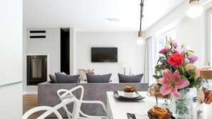 Un salon moderne avec un canapé gris, des chaises blanches, un téléviseur à écran plat et un coin repas avec une table mise pour deux personnes.