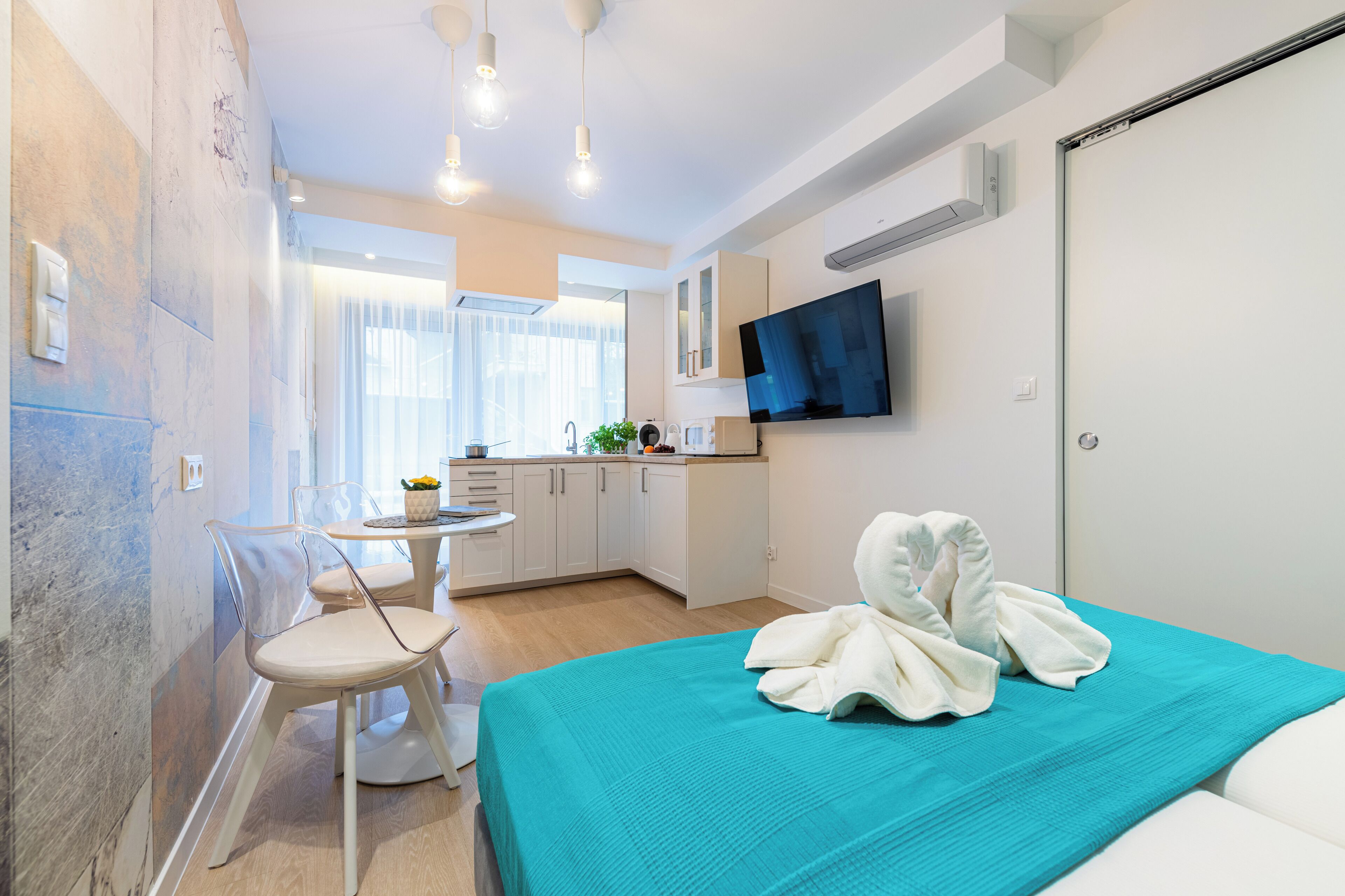 Quarto de hotel moderno com cama, uma mesinha e uma cadeira. Há uma televisão na parede e uma janela com cortinas.