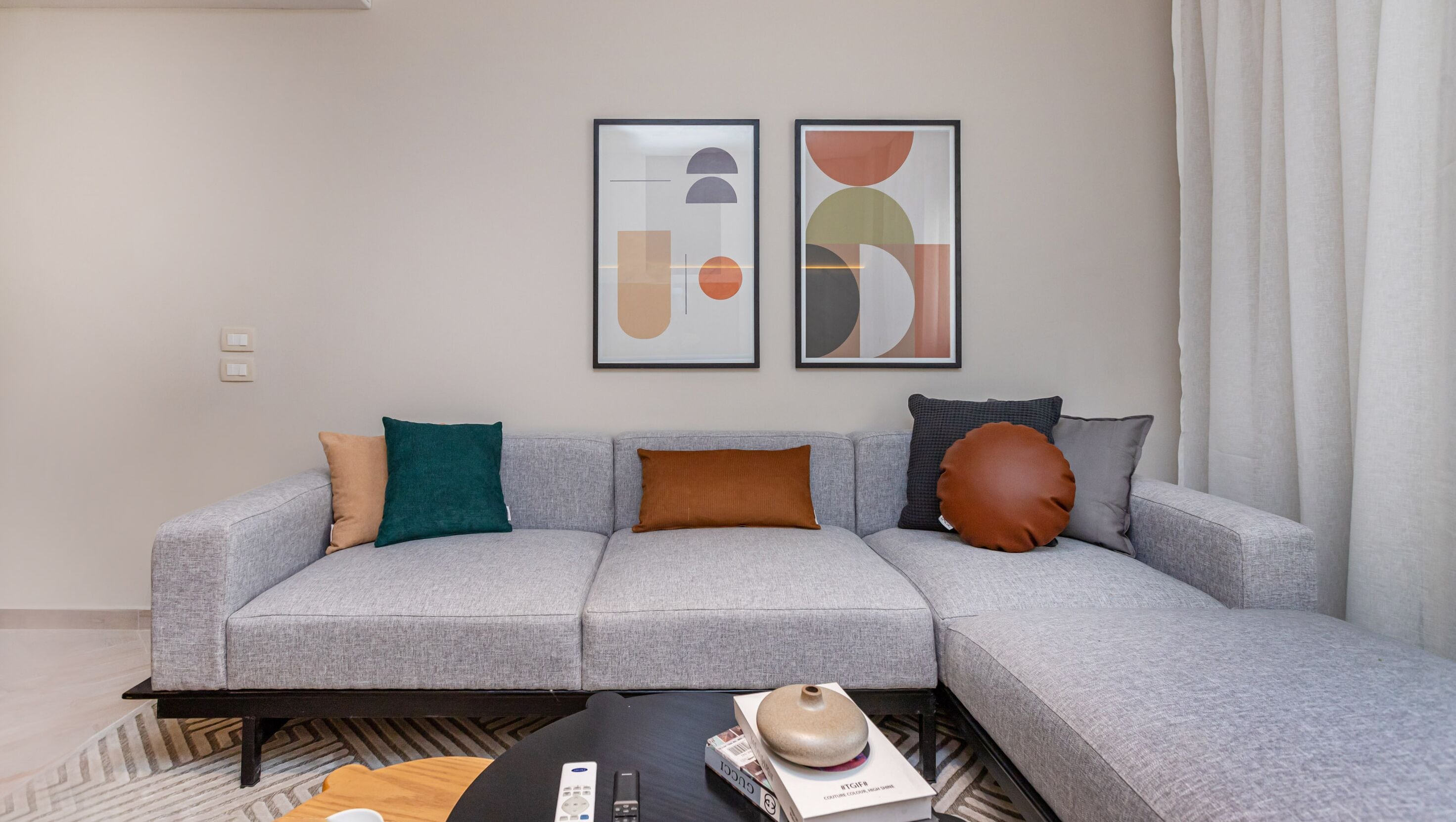 A modern living room with a grey sectional sofa, two framed abstract art pieces, and a coffee table with a book and remote control.
