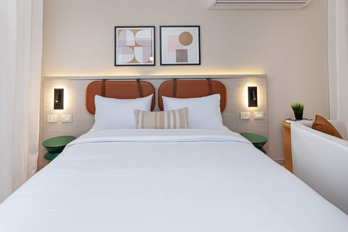 A neatly made bed with white linens, a headboard with a brown leather panel, two wall-mounted lights, and two framed abstract artworks above the bed.