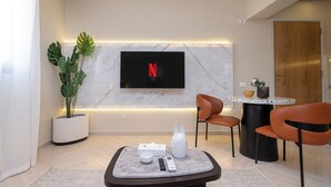 A modern living room with a flat-screen TV mounted on the wall, a potted plant, a coffee table with remote controls, and two chairs. - Prime Residence Point 90 (New Cairo)