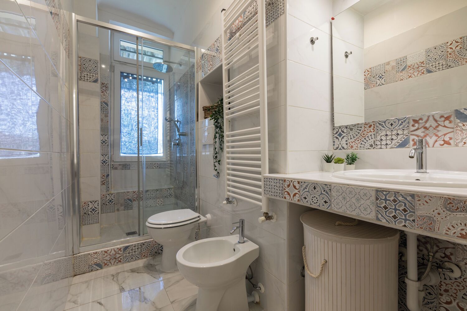 A modern bathroom with a glass-enclosed shower, a toilet, a bidet, and a sink with a decorative tile backsplash.