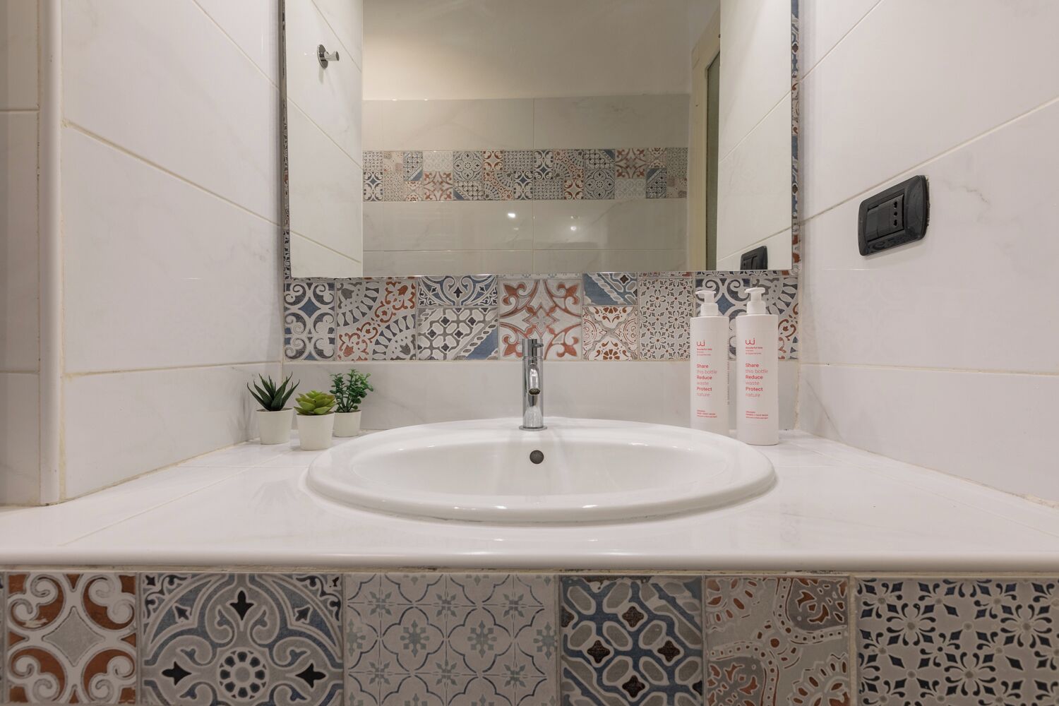 A bathroom with a white sink, a mirror, and a tiled backsplash. There are two small potted plants on the counter.