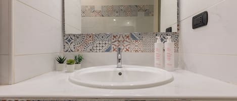 A bathroom with a white sink, a mirror, and a tiled backsplash. There are two small potted plants on the counter.