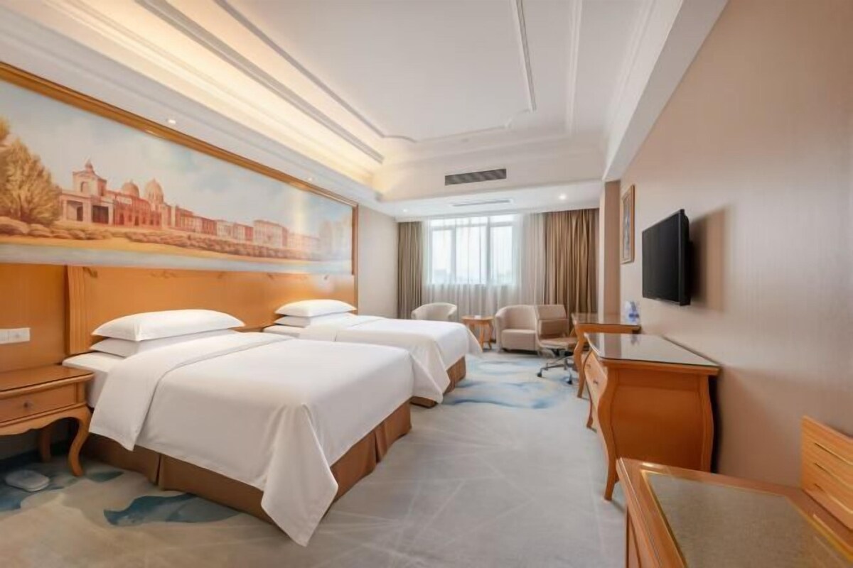 A hotel room with two beds, a desk, a chair, a flat-screen TV, and a large window.