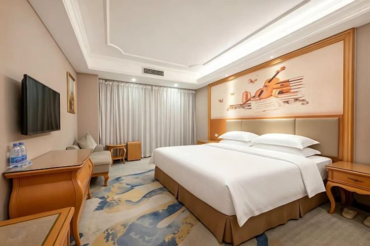 A hotel room with a bed, bedside tables, a TV, a desk, and a large window with curtains.