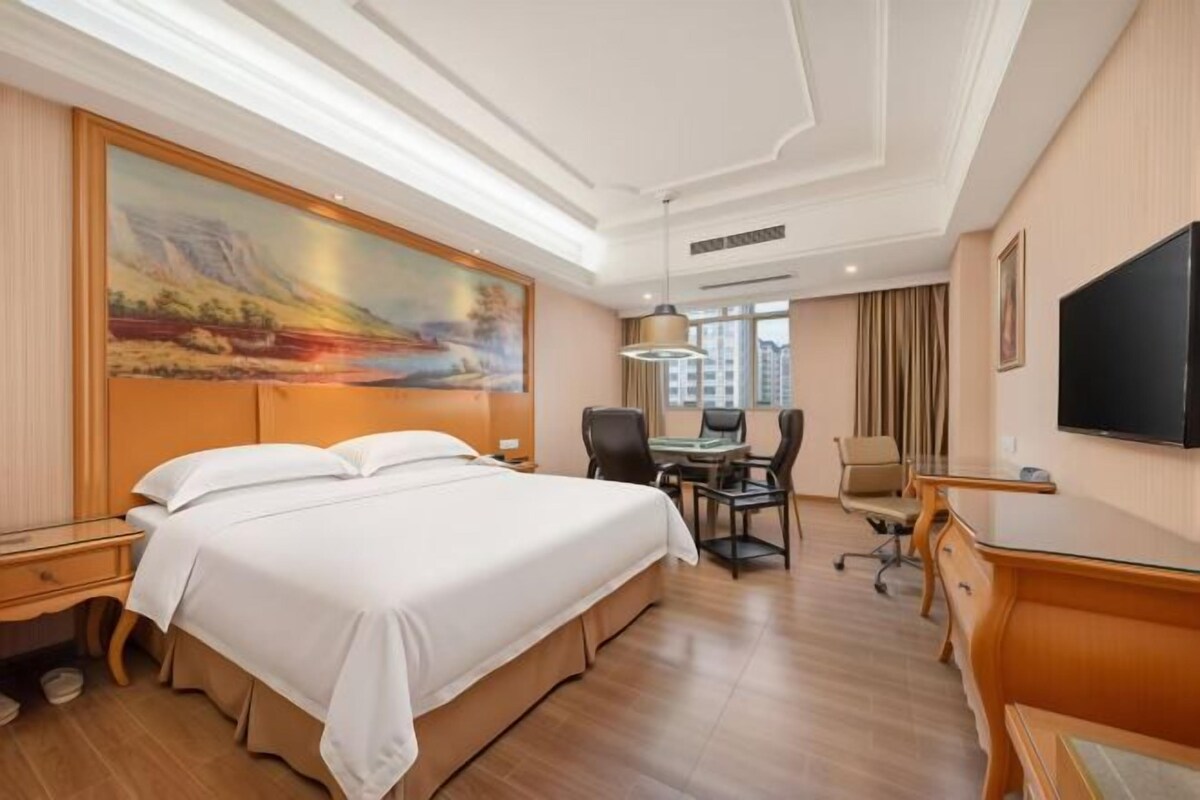 A hotel room with a large bed, a desk, a chair, a television, and a painting on the wall.
