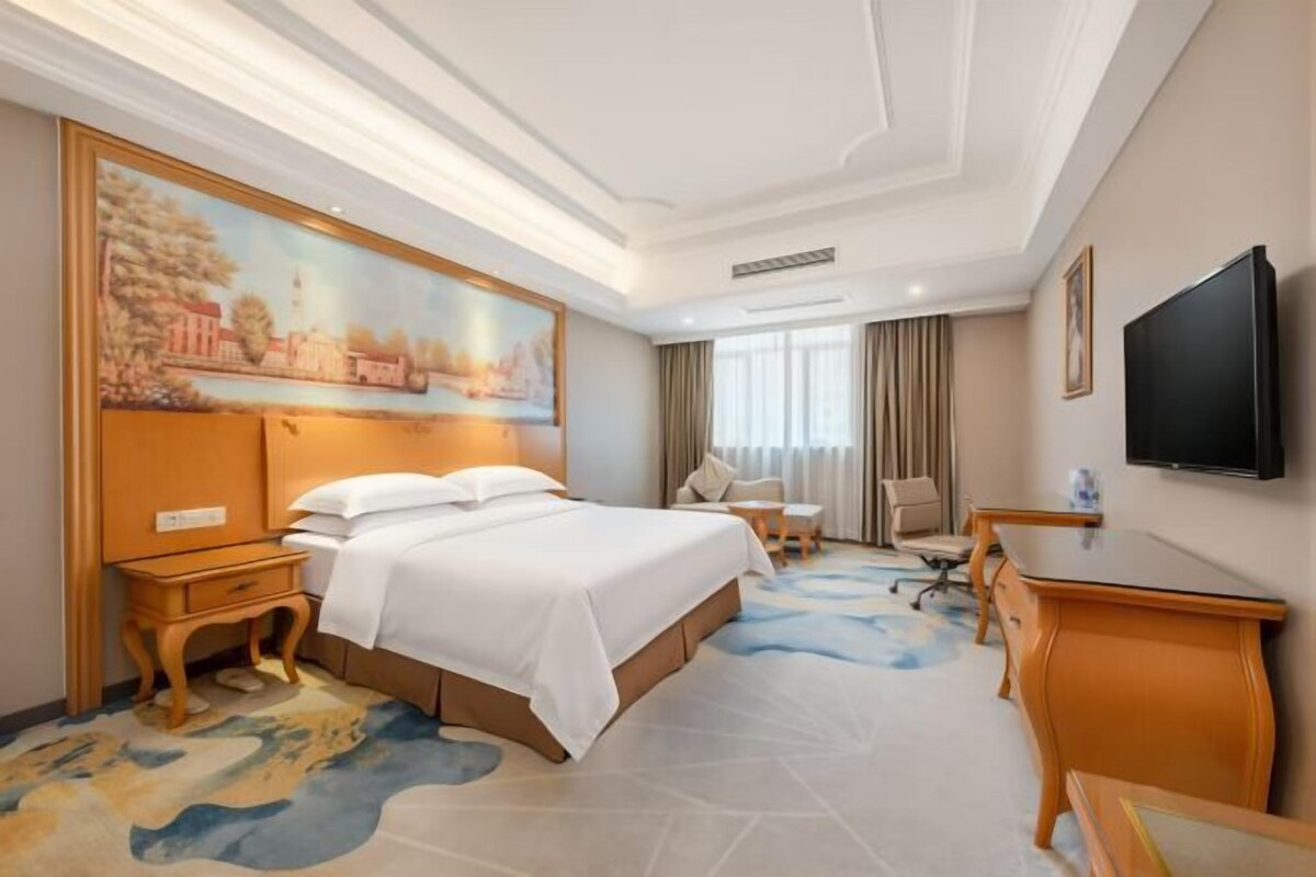 A hotel room with a large bed, a desk, a chair, a television, and a painting on the wall.